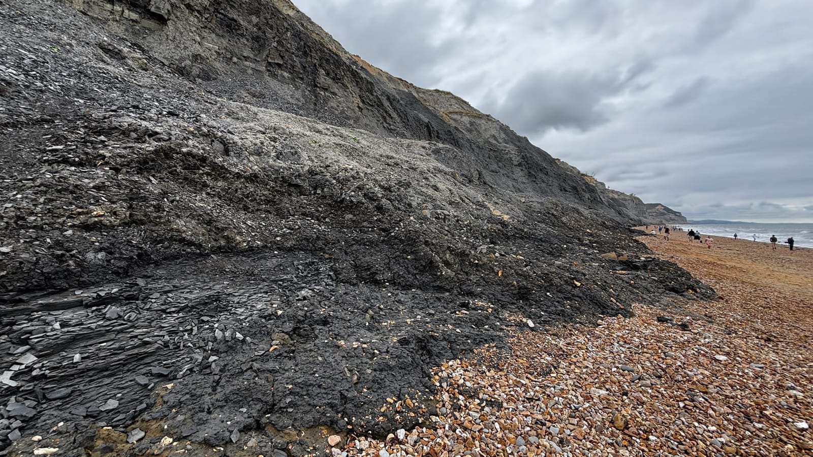 Dark grey clay cliffs above a shingle beach at Charmouth, with fossil hunters along the shoreline